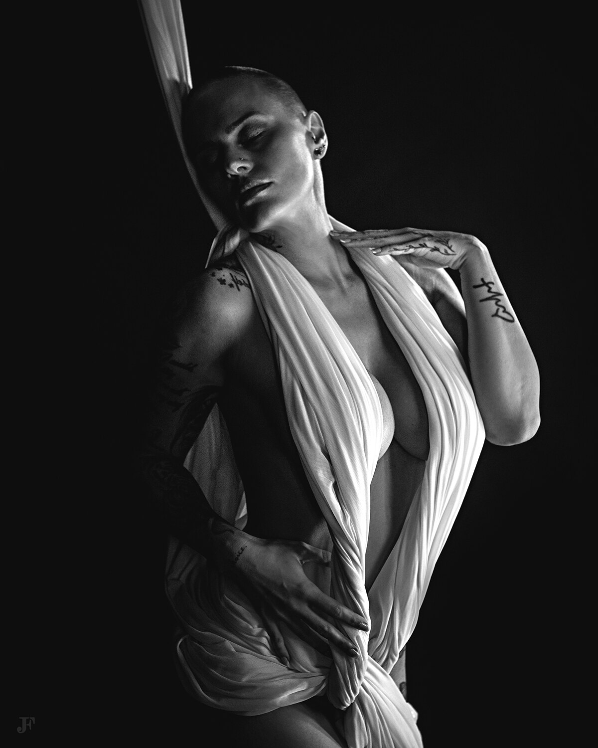 The image shows a black and white photo of a woman with tattoos on her arms, standing in a dark background. She is wearing a cloth draped over her body, and there is a watermark in the bottom left corner of the image.
