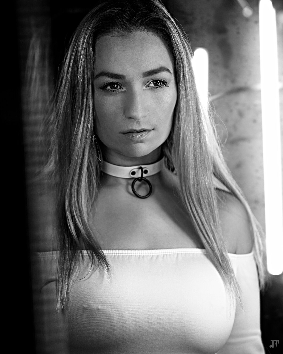 The image shows a woman with long blonde hair wearing a white dress and a choker, standing in front of a wall with lights in the background.