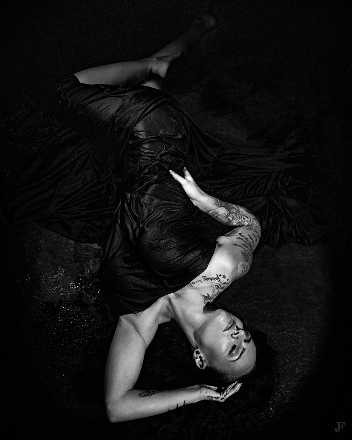 The image shows a woman in a black dress laying on the ground with her arms outstretched, her face illuminated by the darkness of the background.