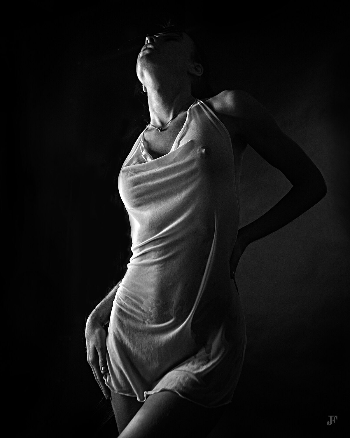 The image shows a woman in a white dress standing in the dark. She is facing away from the viewer, her arms outstretched and her head tilted slightly to the side. Her hair is pulled back in a bun and she has a gentle expression on her face. The darkness o