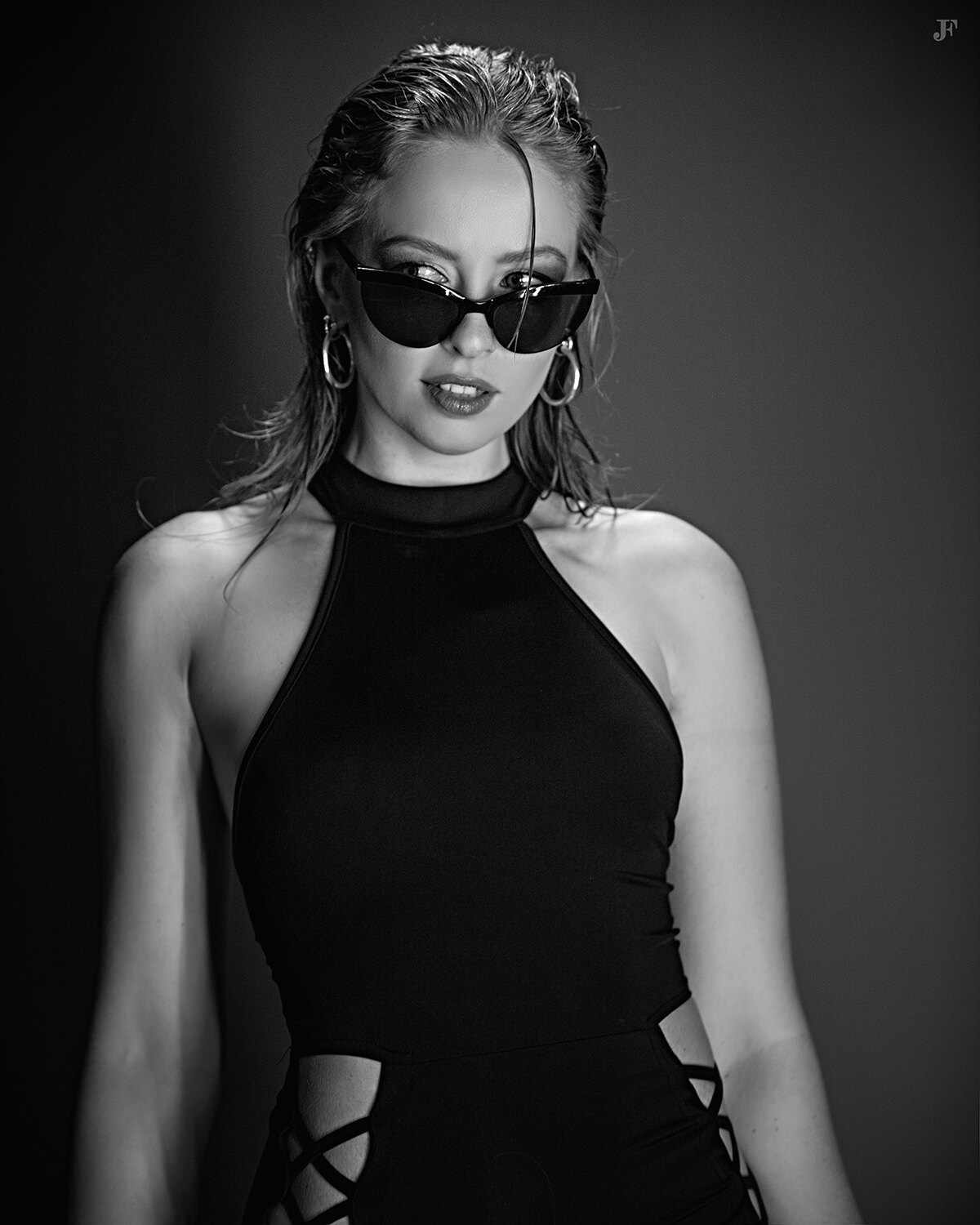 The image shows a woman in a black dress and sunglasses posing for a picture. She is wearing a pair of sunglasses and her hair is styled in a sleek, straight look. Her makeup is subtle and her expression is confident and poised.