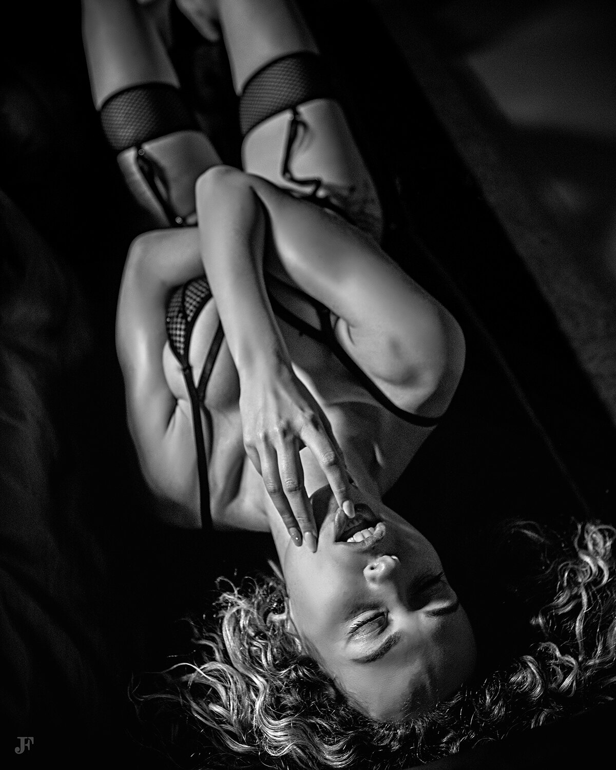 The image shows a woman laying on top of a bed in a black and white setting. She is wearing lingerie and her hair is styled in a sleek bob. Her eyes are closed and her lips are slightly parted, giving her a peaceful and serene look.
