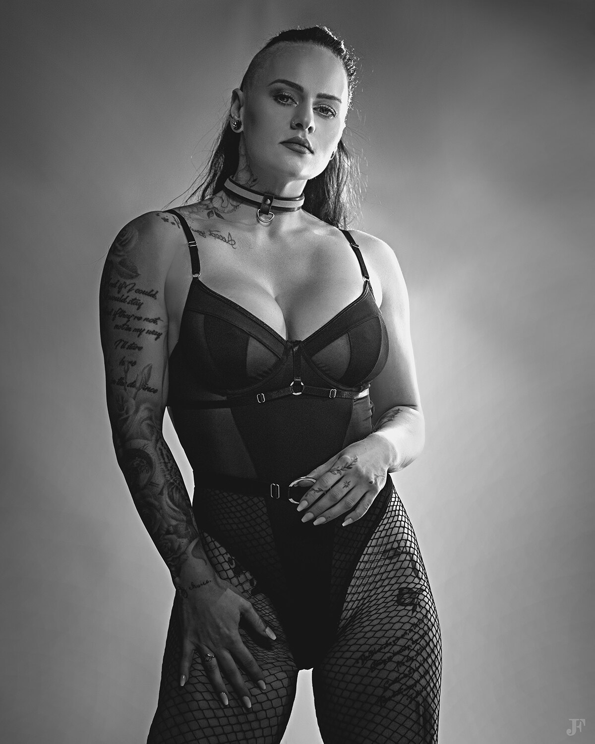 The image shows a woman in a black bodysuit and fishnet stockings posing for a picture. She has tattoos on her body and there is a watermark in the bottom right corner of the image.
