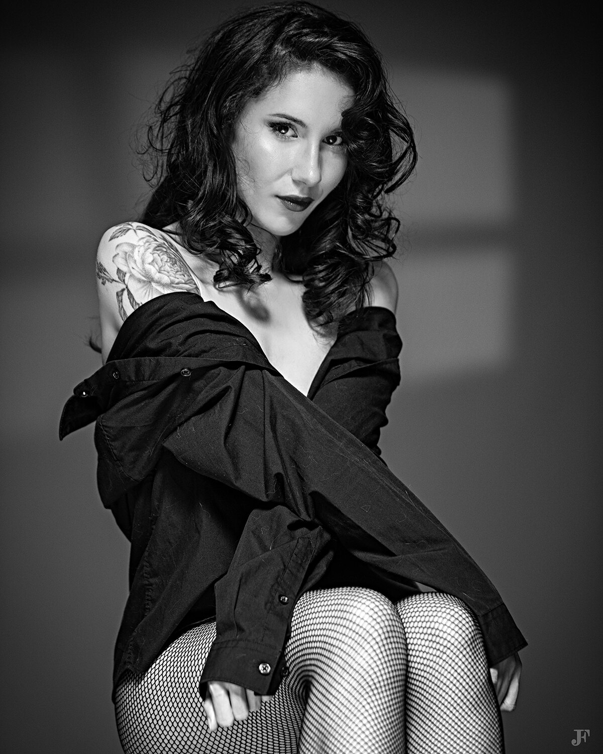 The image shows a woman in fishnet stockings posing for a black and white photo. She is wearing a shirt and has a tattoo on her arm.