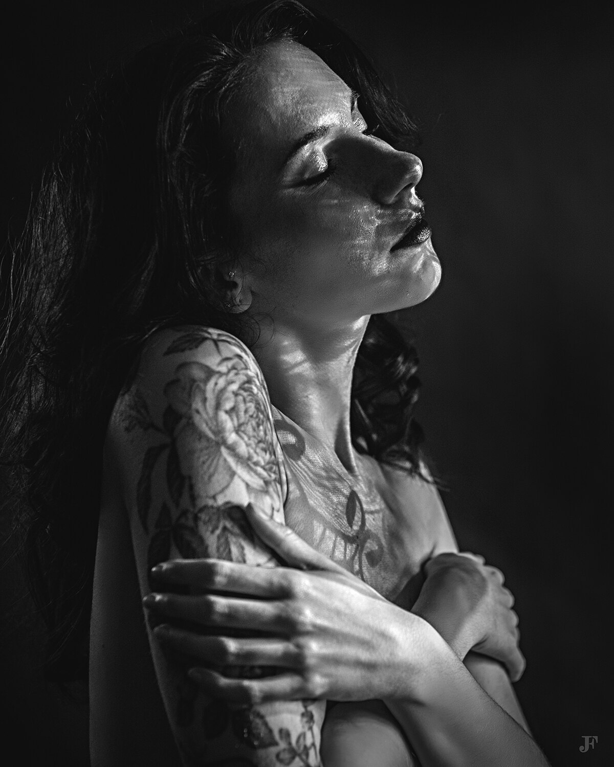 The image shows a woman with long hair and tattoos on her arms against a dark background. The image is in black and white, giving it a timeless and classic feel.