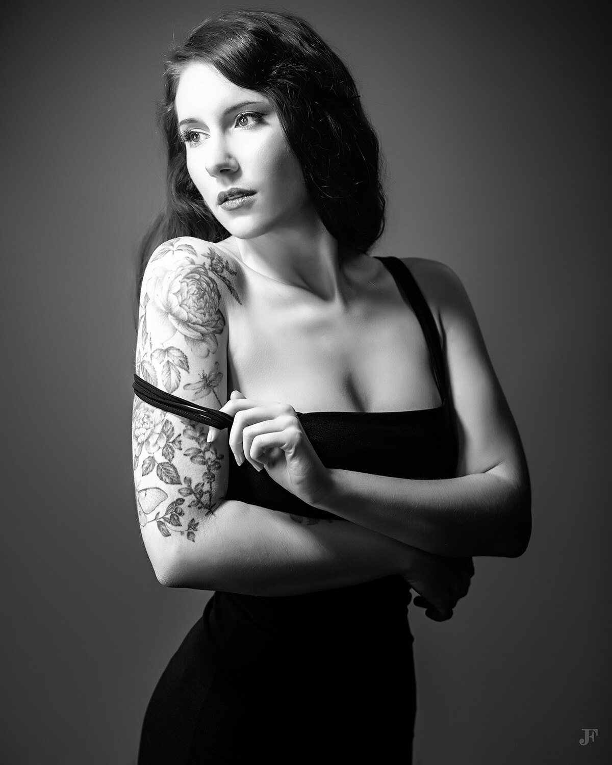 The image shows a woman in a black dress with a tattoo on her arm. She has a serious expression on her face, and her hair is pulled back in a neat bun. The tattoo is intricate and detailed, with a variety of colors and shapes.