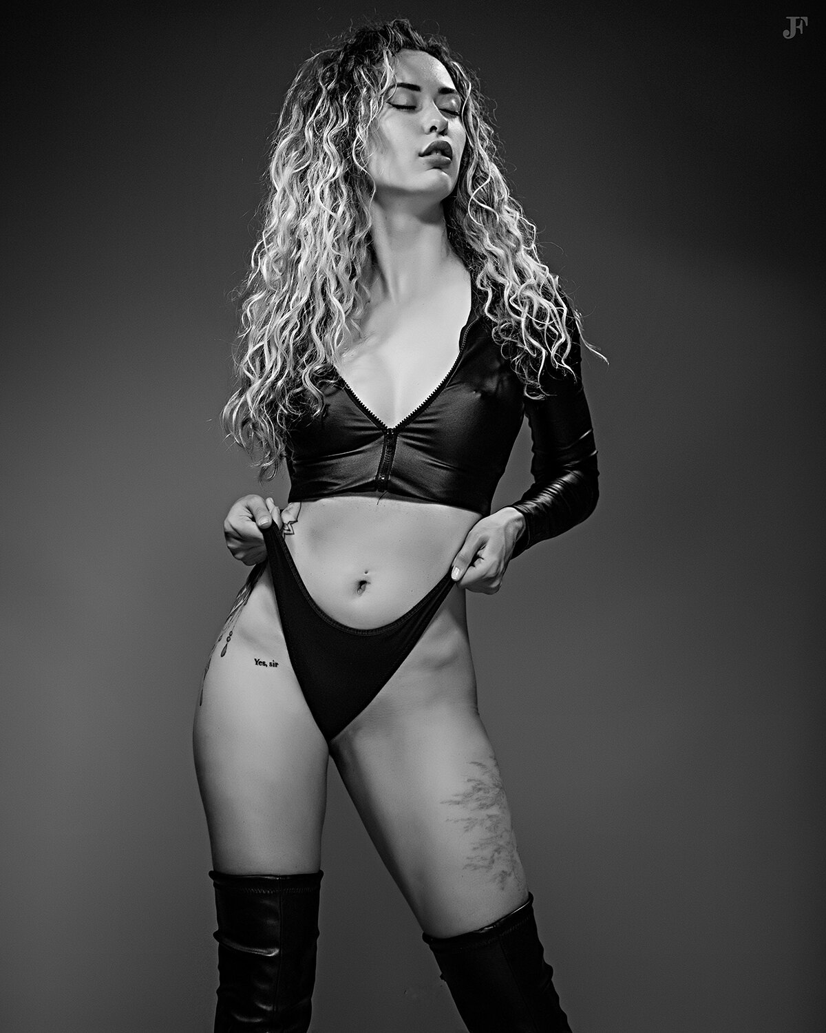 The image shows a woman in a black bikini and thigh high boots posing for a black and white photo. She is standing in the center of the frame, her body illuminated by the light of the setting sun. Her hair is styled in a sleek bob and her makeup is subtle