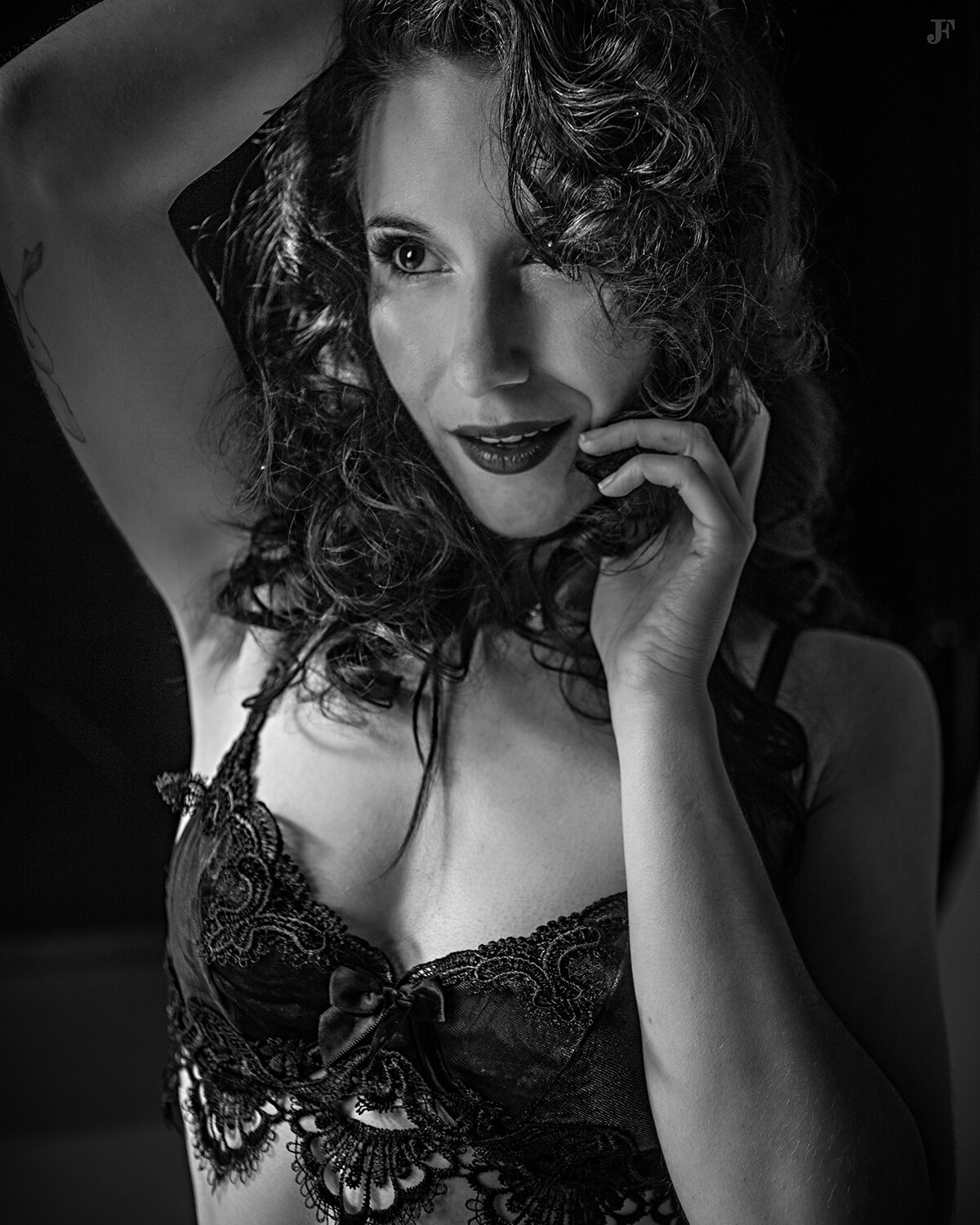 The image shows a woman in a black lingerie posing for a picture against a dark background. Her face is clearly visible, with her eyes looking directly at the camera and her lips slightly parted. Her hair is pulled back in a neat bun, and her skin is glow