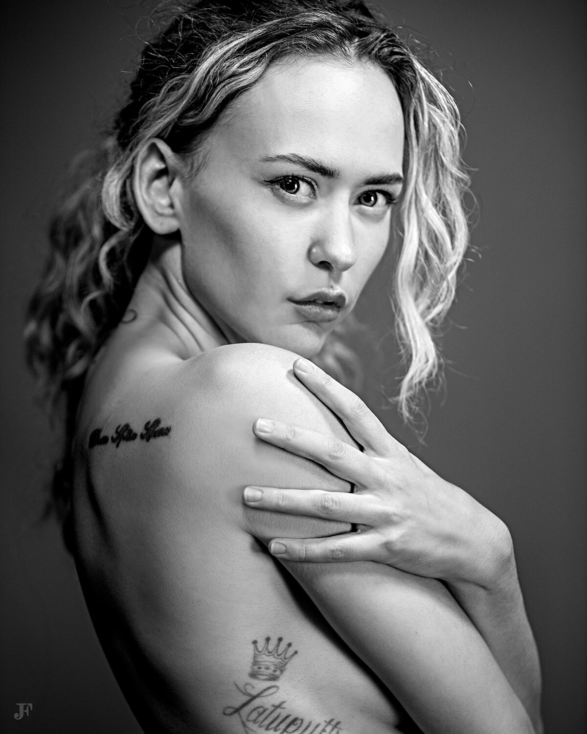 The image shows a black and white portrait of a woman with a tattoo on her back. The tattoo is intricate and detailed, with a variety of shapes and symbols. The woman has a serious expression on her face, and her hair is pulled back in a neat bun. The bac