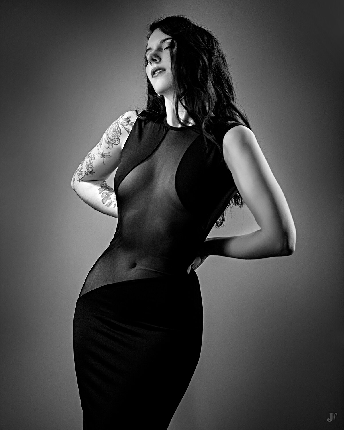 The image shows a woman in a black dress posing for a picture. She has a tattoo on her arm, and the image is in black and white.