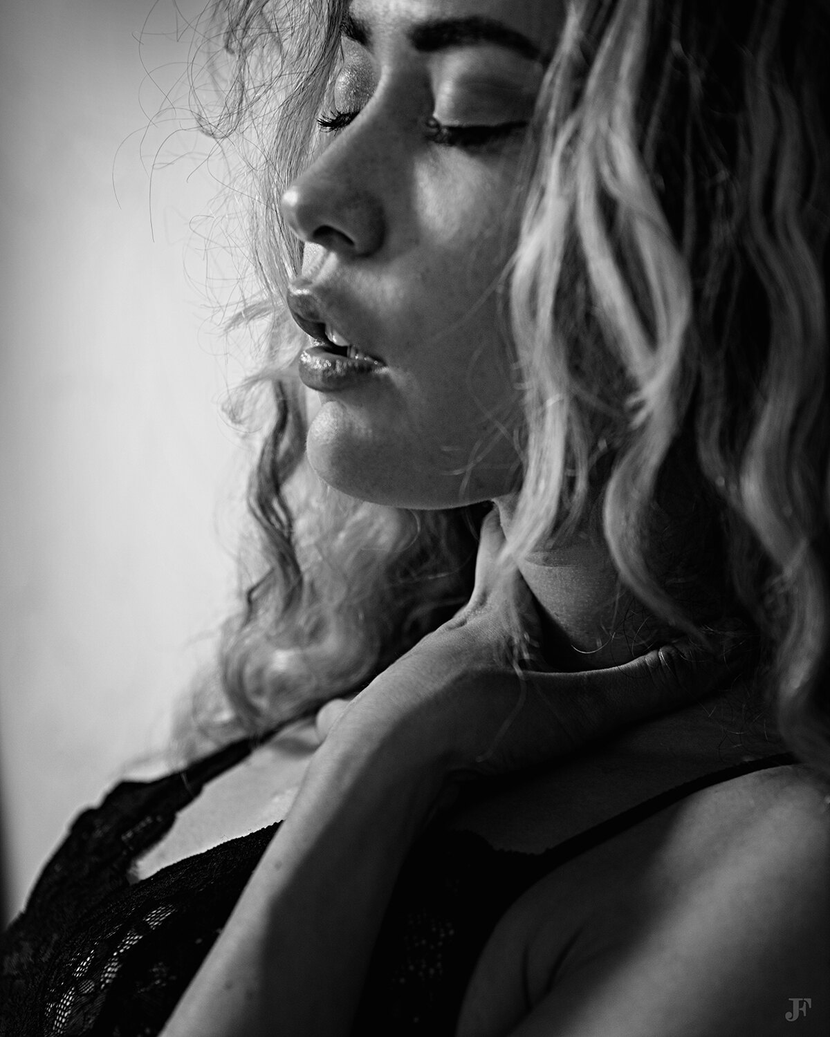 The image shows a woman with long blonde hair wearing a black dress, standing in front of a wall. Her face is clearly visible, with her eyes closed and her lips slightly parted.