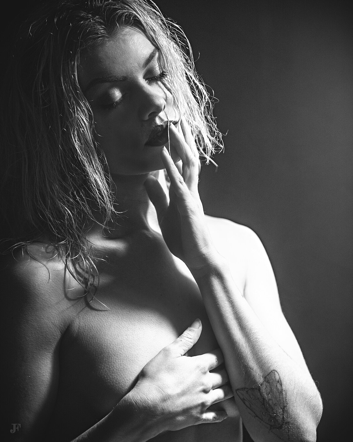 The image shows a black and white photo of a woman with long hair and a tattoo on her arm. She is standing in the center of the frame, her hair cascading down her back and her eyes closed, giving her a peaceful and serene look.