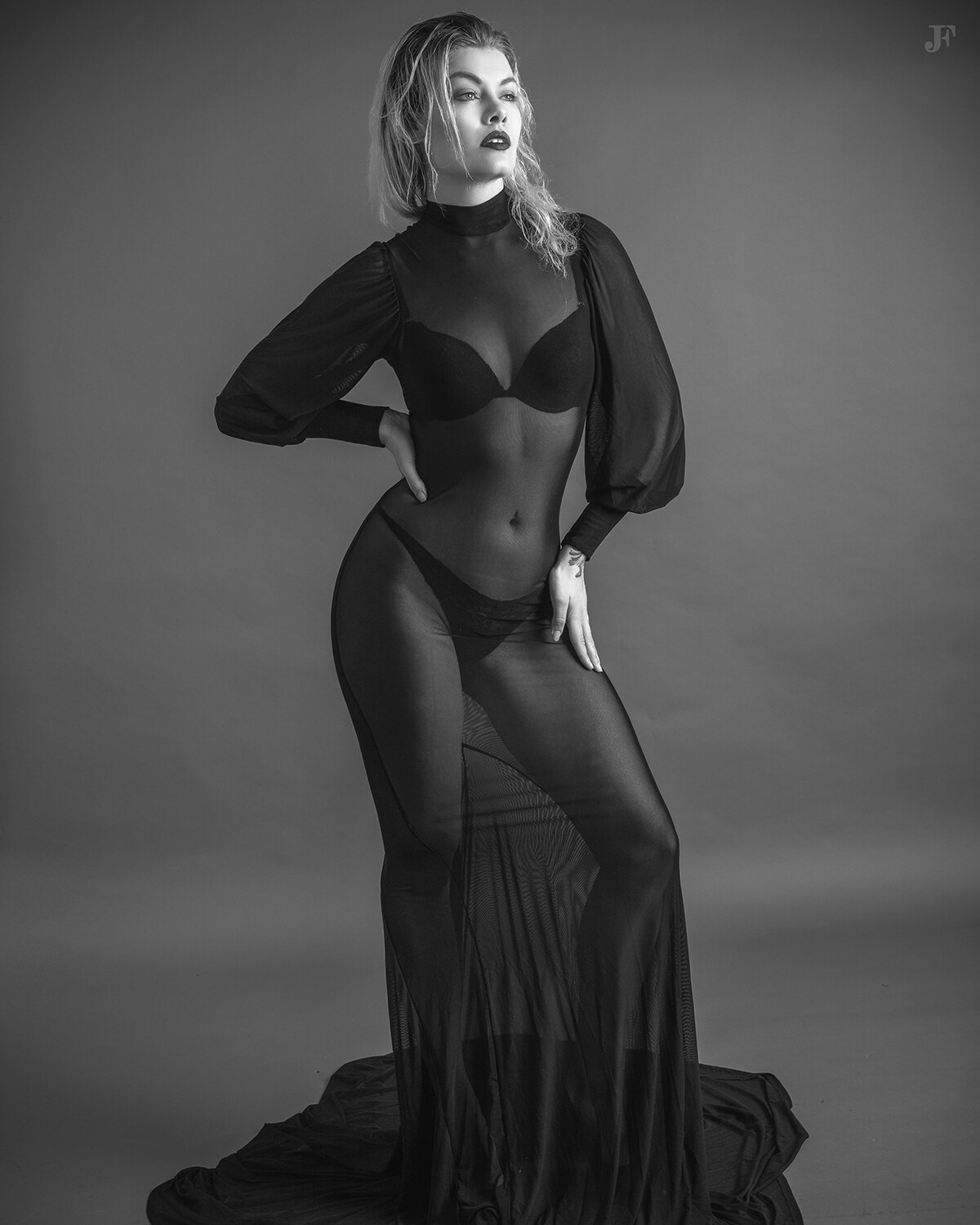 The image shows a woman in a black dress posing for a black and white photo. She is standing in the center of the frame, her dress billowing around her as she poses for the camera. Her hair is styled in a sleek bob and her makeup is subtle and natural. Sh