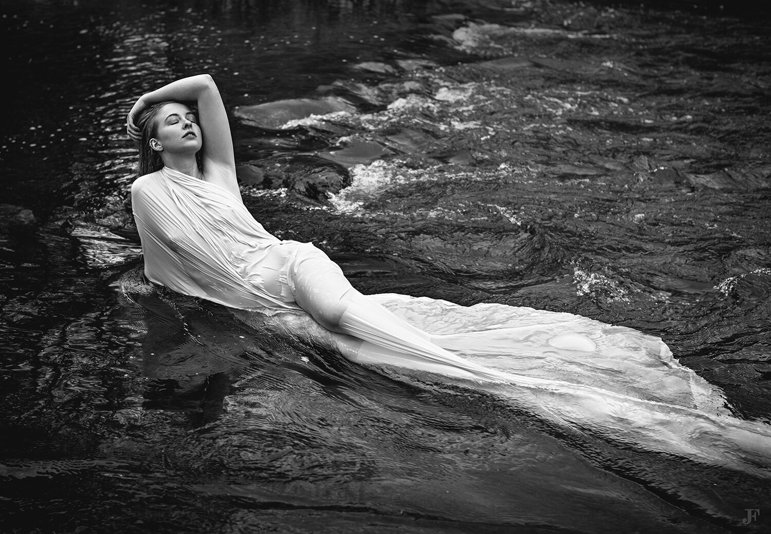 The image shows a woman in a white dress floating in the water, captured in a black and white photography session. The woman is surrounded by rocks, creating a peaceful atmosphere.