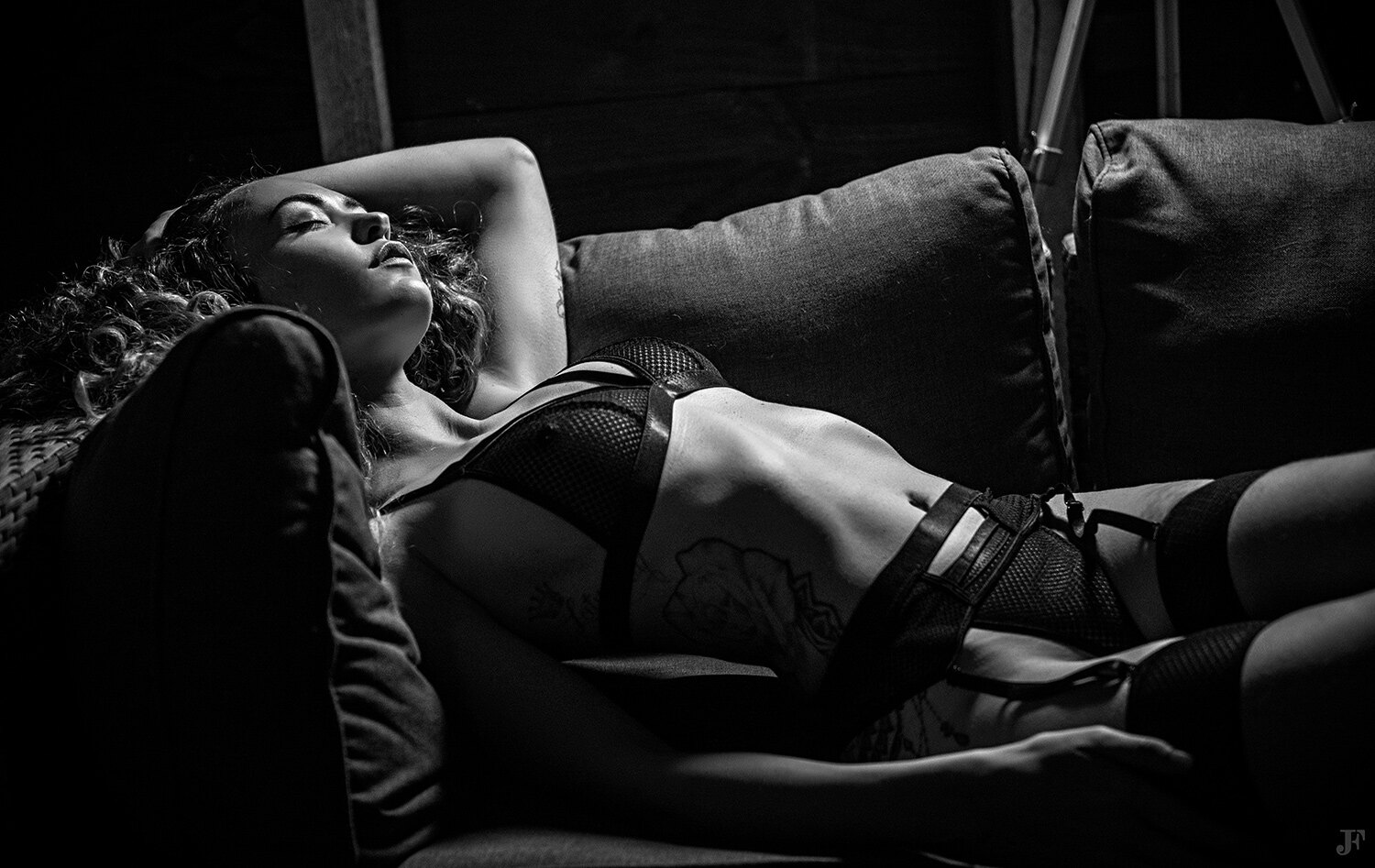 The image shows a woman laying on top of a couch in a black and white setting. She is surrounded by pillows and there is a wall in the background.