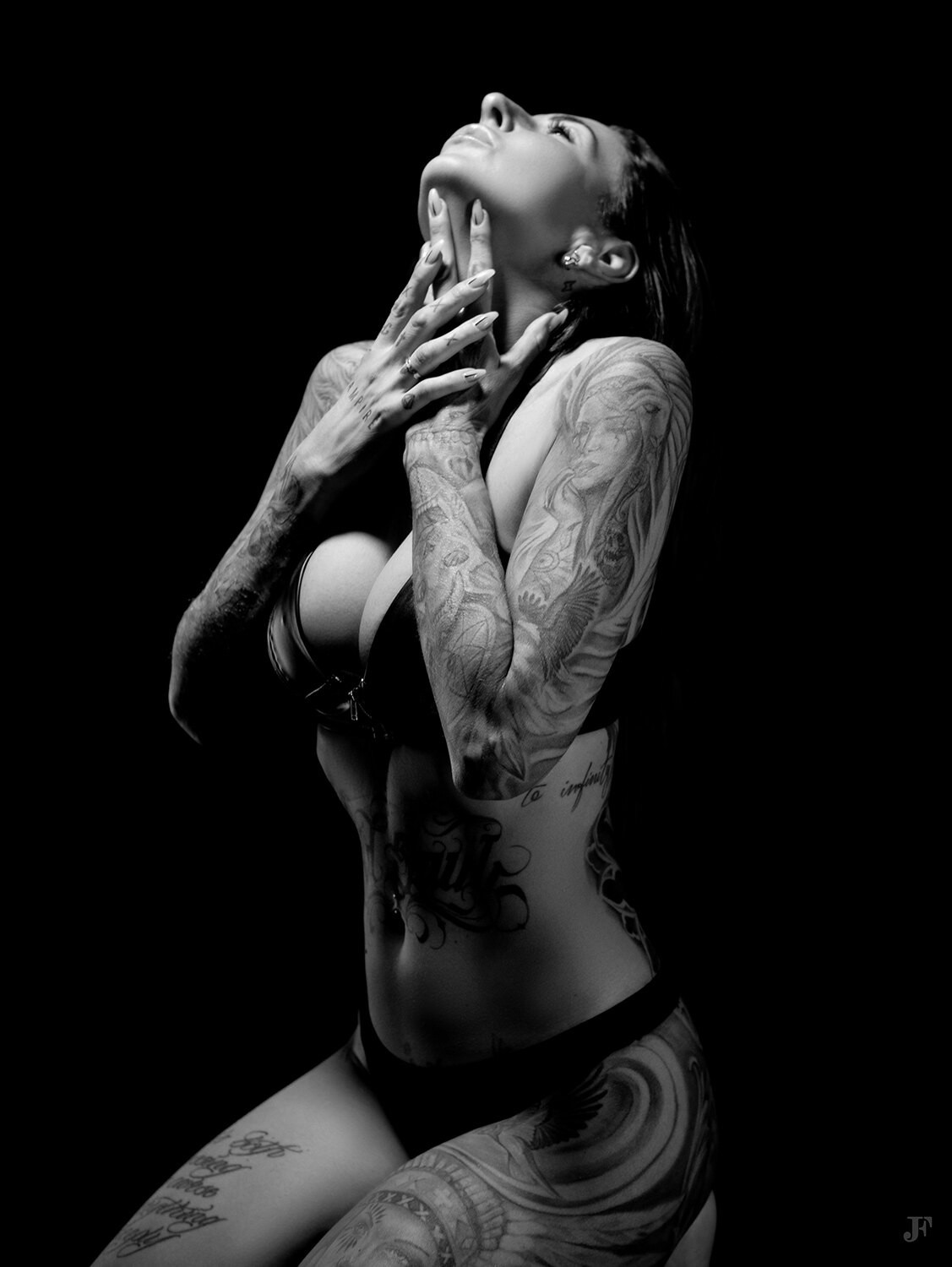 The image shows a woman with tattoos on her body posing for a black and white photo against a dark background.