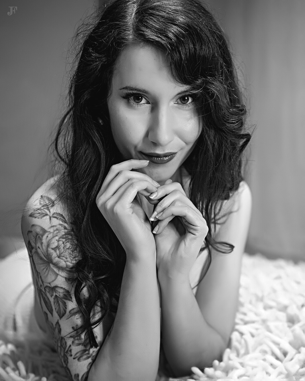 The image shows a woman laying on top of a white blanket, with a tattoo on her arm. The background is blurred, and there is a watermark on the image.