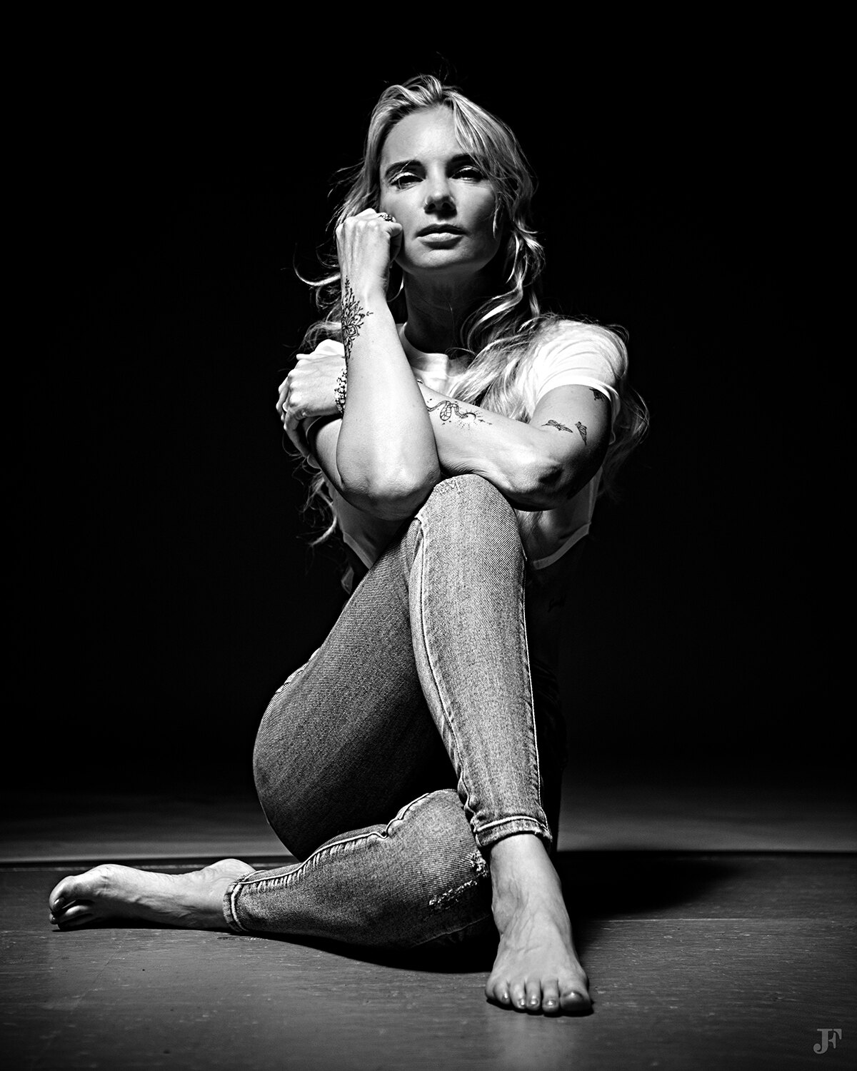 The image shows a woman sitting on the floor with her legs crossed, wearing a white t-shirt and jeans. The background is black and there is a watermark in the bottom right corner of the image.