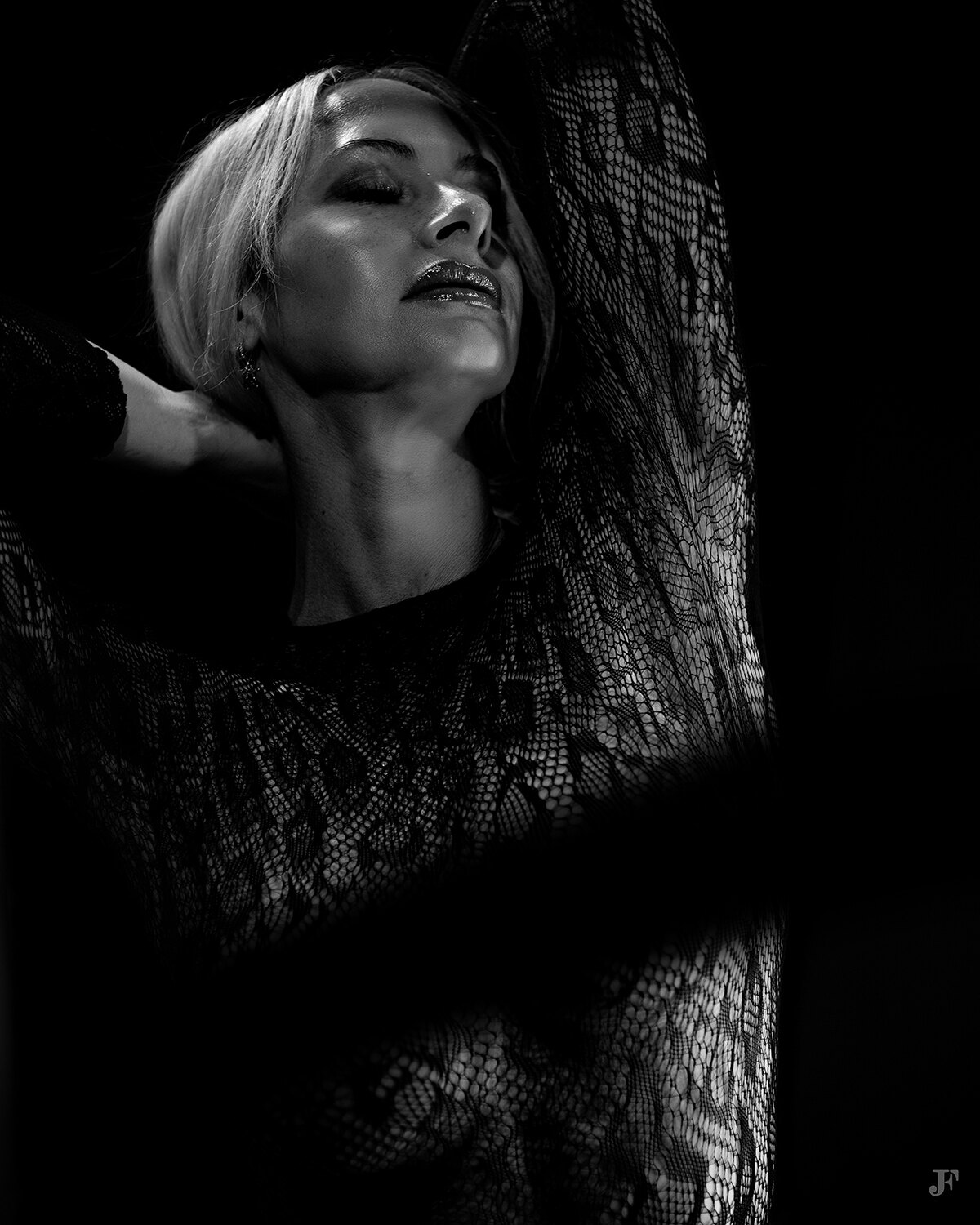 The image shows a woman in a black dress with her arms outstretched, standing in a dark background. She appears to be in a trance-like state, with her eyes closed and her hands clasped together in front of her. Her hair is pulled back in a bun and her face is illuminated by a soft light, giving her a mysterious and captivating look.